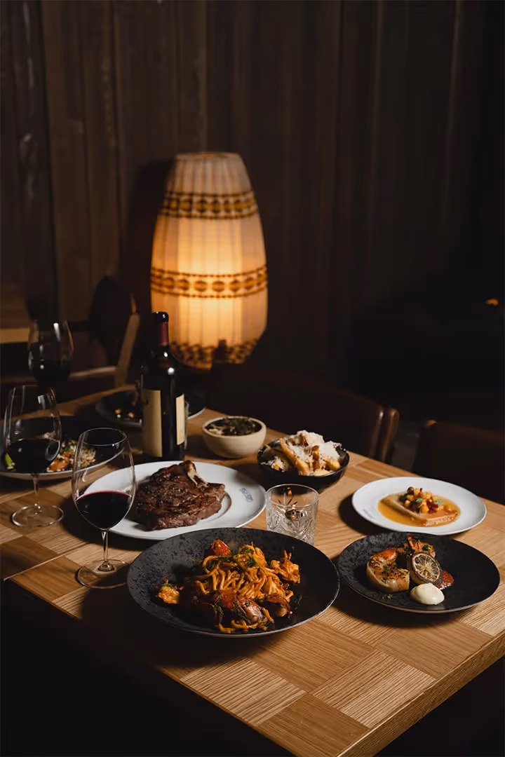 Restaurant table with steak, pasta, seafood dishes, wine glasses, and a bottle under warm lighting.