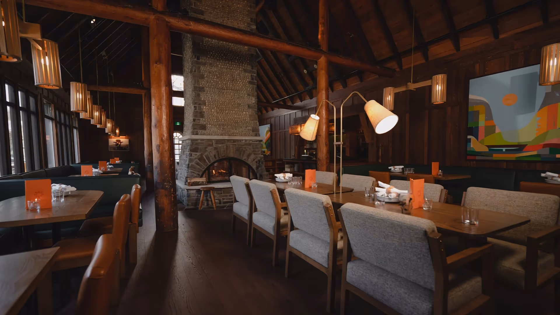 Rustic restaurant interior with wooden beams, stone fireplace, and tables with chairs and lamps.