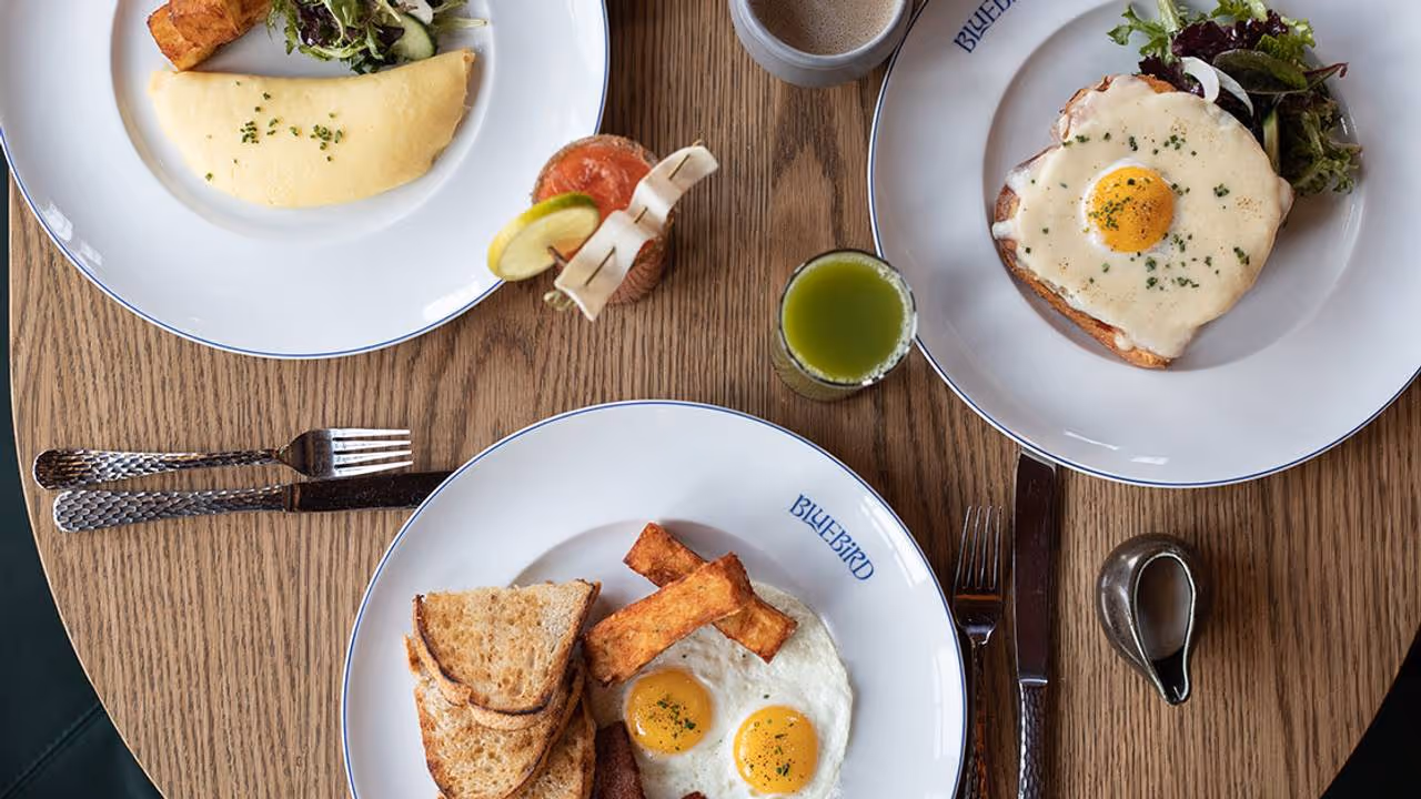 Table with three breakfast plates including eggs, toast, omelette, salad, and drinks on a wooden surface.