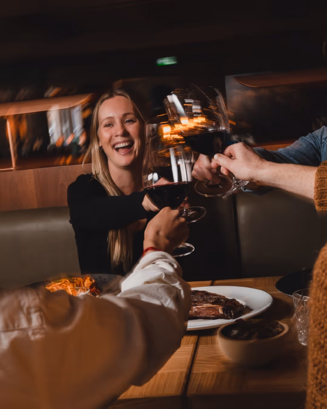 Smiling woman and friends clinking glasses of red wine over a dinner table with steak and pasta.