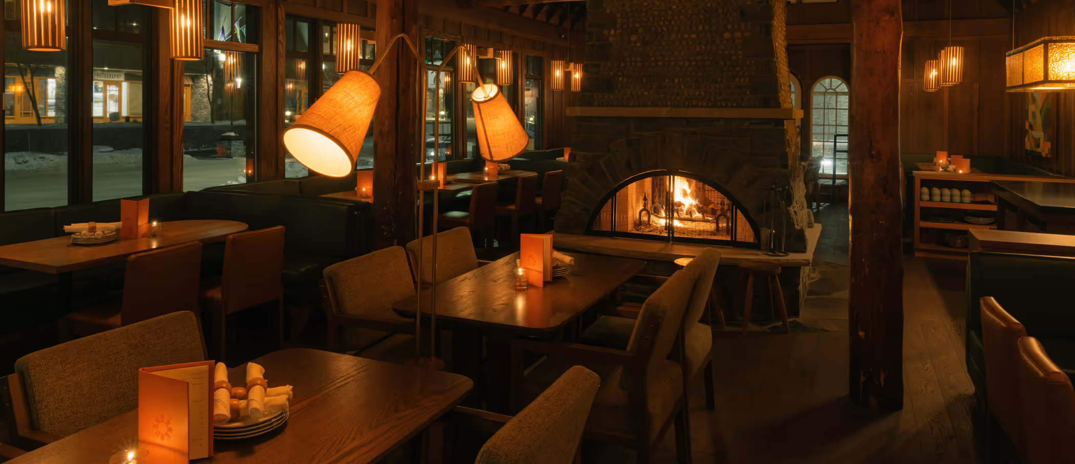 Cozy restaurant interior with wooden tables, upholstered chairs, warm lamps, and a stone fireplace with a fire.