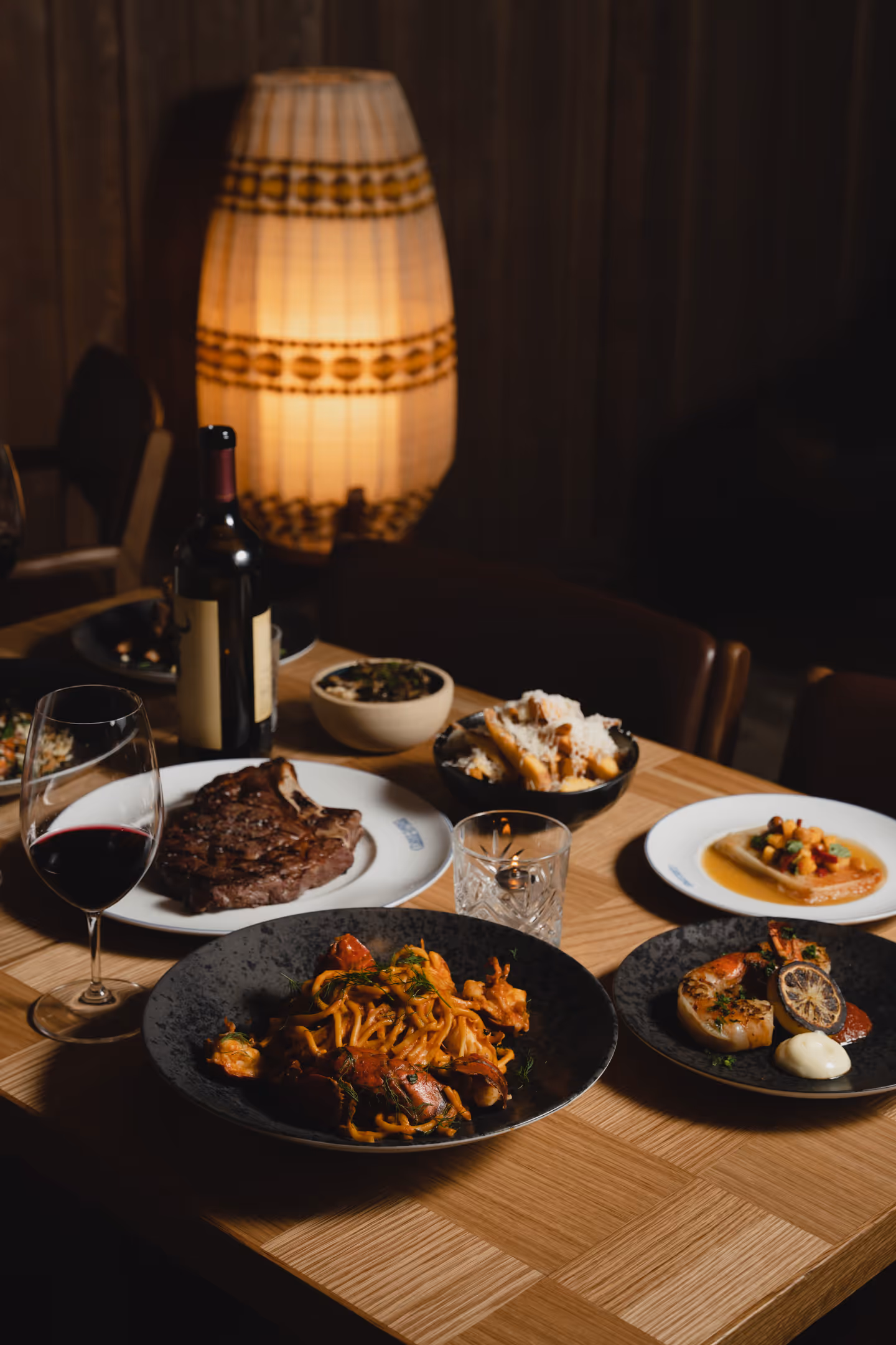 Dining table with steak, seafood pasta, grilled shrimp, fries, wine, and a lit lamp in background.