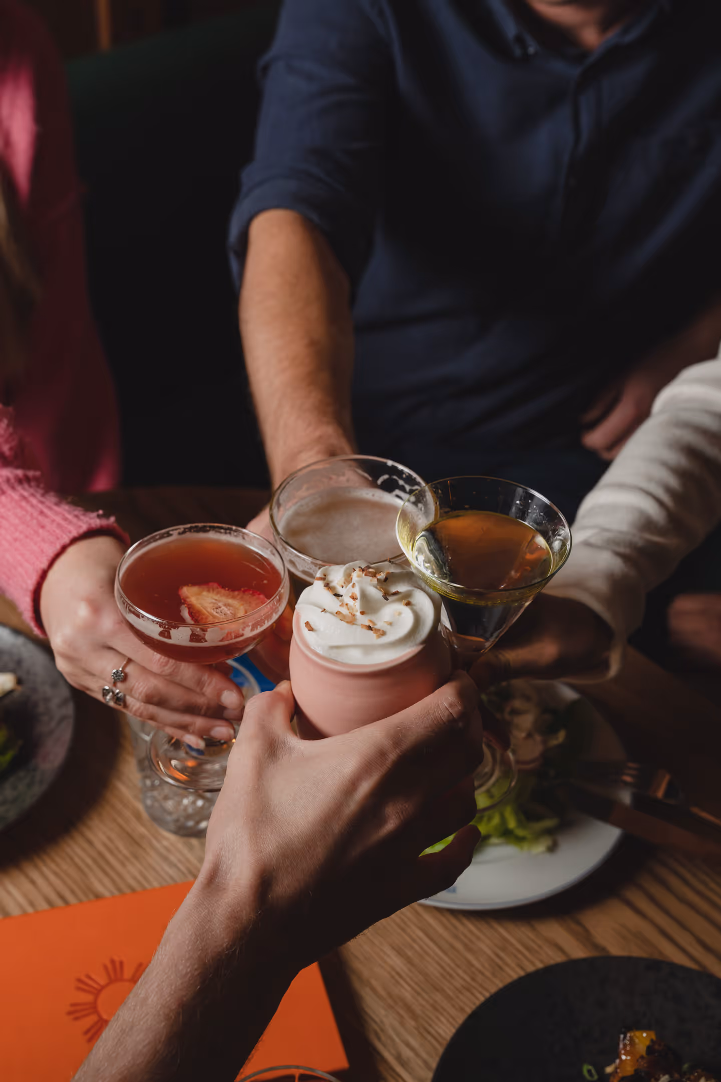 Four people clinking different cocktails and drinks over a table with plates of food.