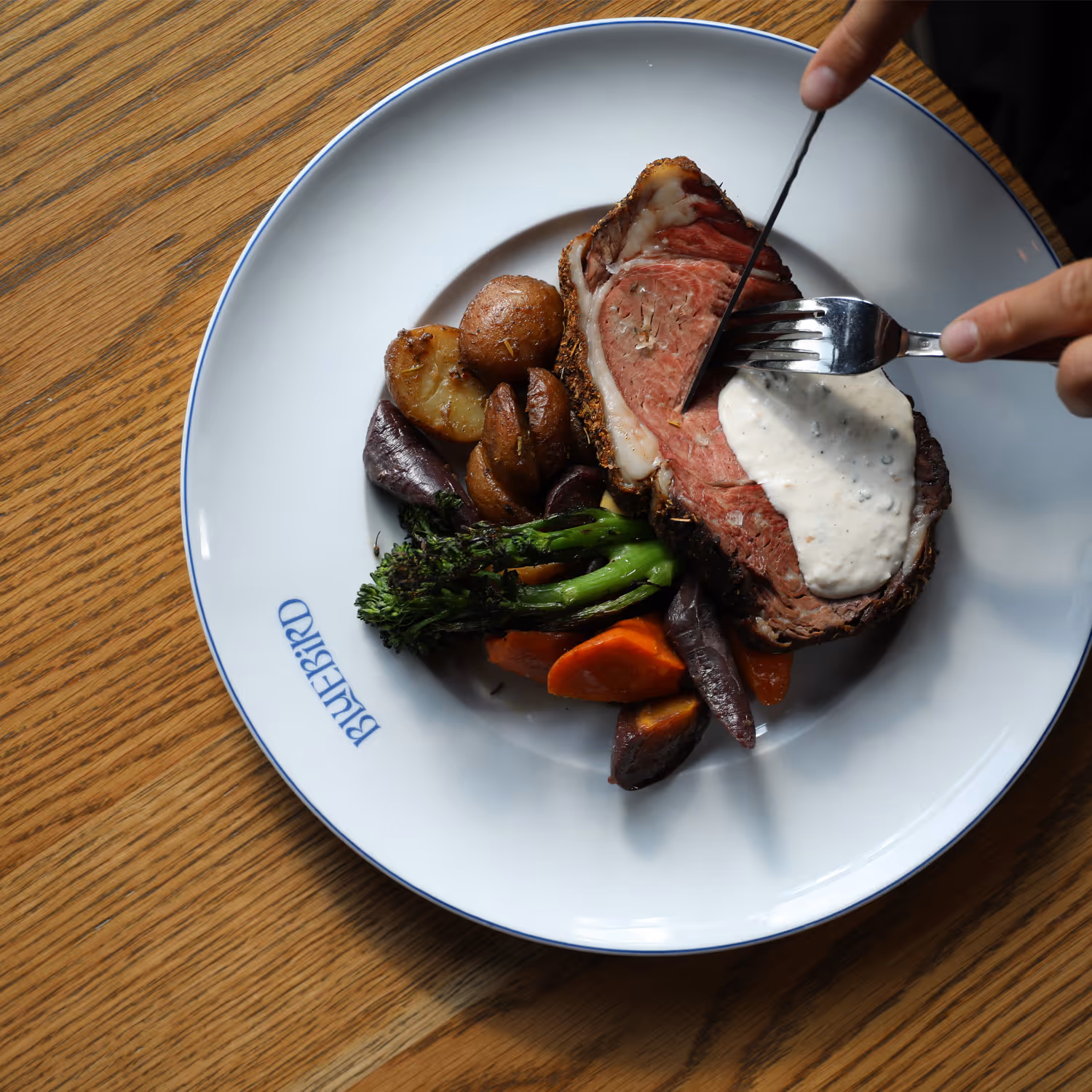 Plate of sliced roast beef with horseradish sauce, roasted potatoes, carrots, and broccolini on a wooden table.