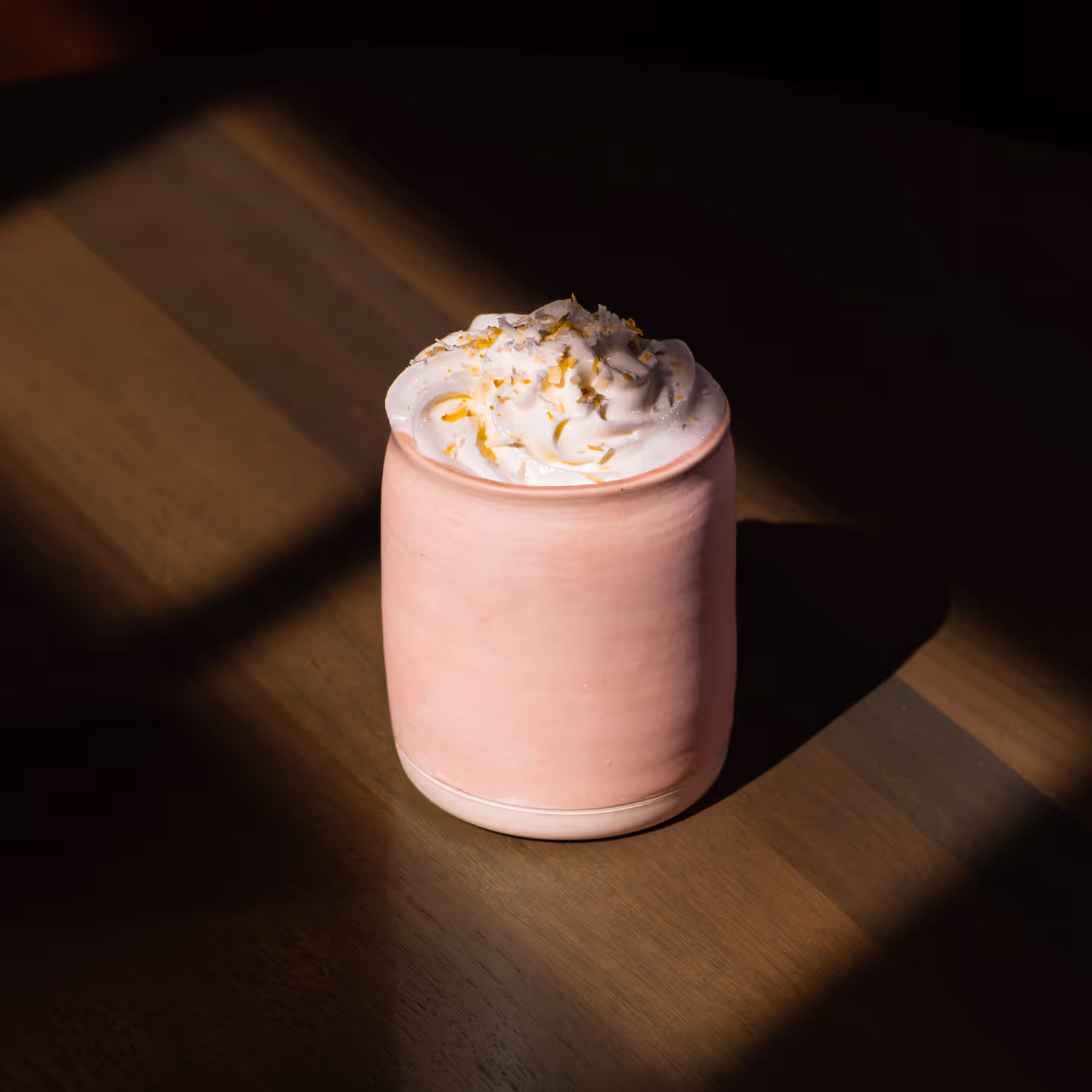 Pink ceramic mug with whipped cream and orange zest on a wooden table with light and shadow.