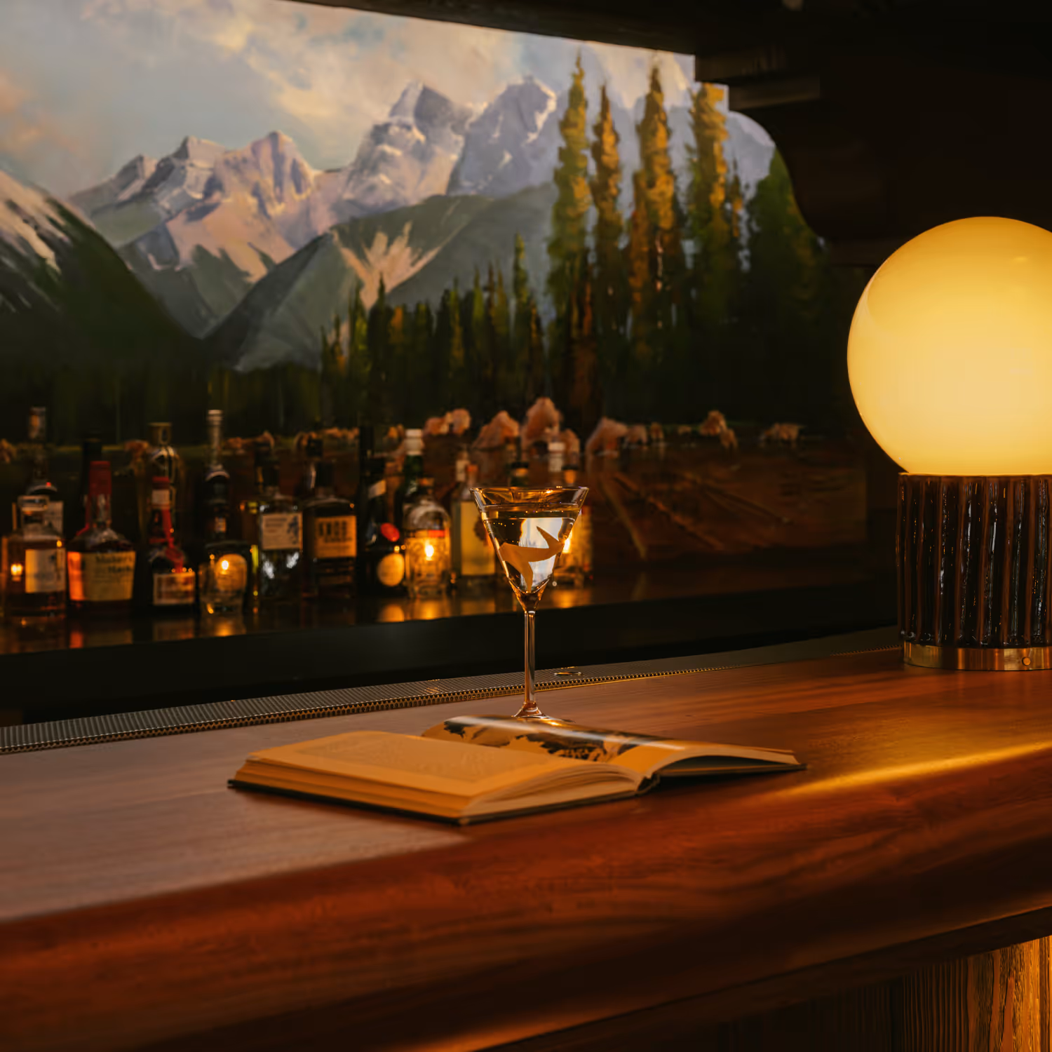 Martini glass and open book on a wooden bar counter with a glowing round lamp and mountain mural background.