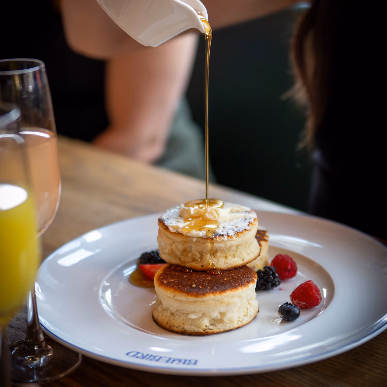 Maple syrup being poured over thick stacked pancakes with butter and mixed berries on a white plate.