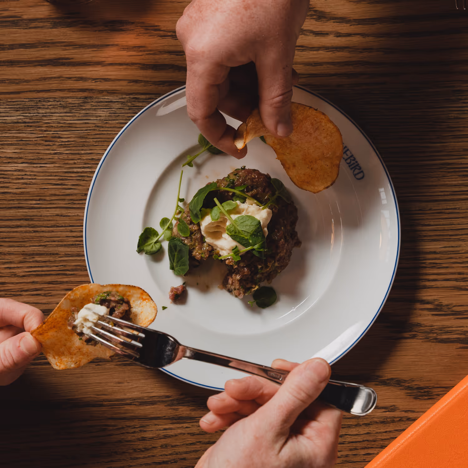 Hands serving beef tartare with cream and greens on a potato chip over a white plate on a wooden table.