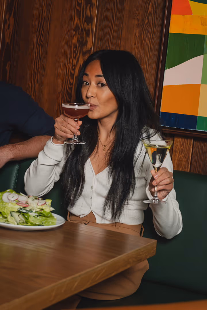 Woman with long black hair holding and sipping from two cocktail glasses at a wooden table with a salad plate nearby.