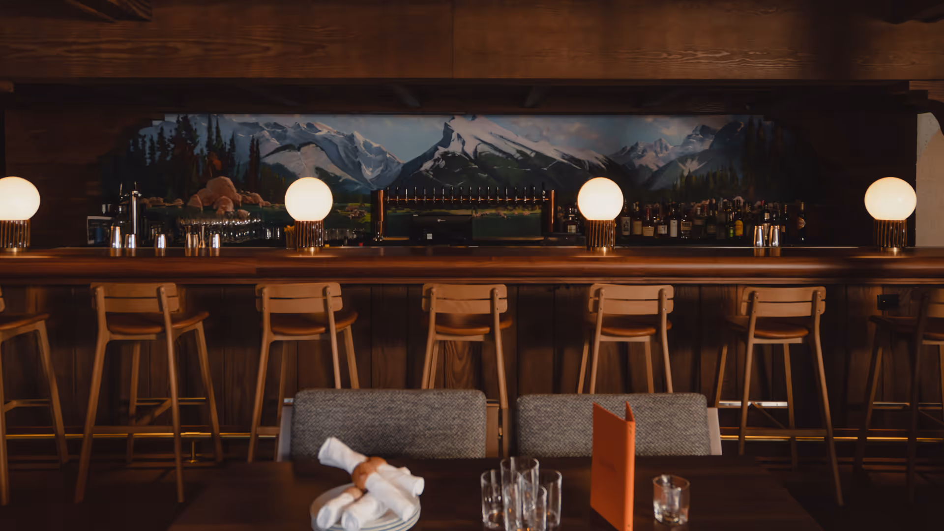 Rustic bar with wooden stools, round lamps, and a mountain mural behind the counter.