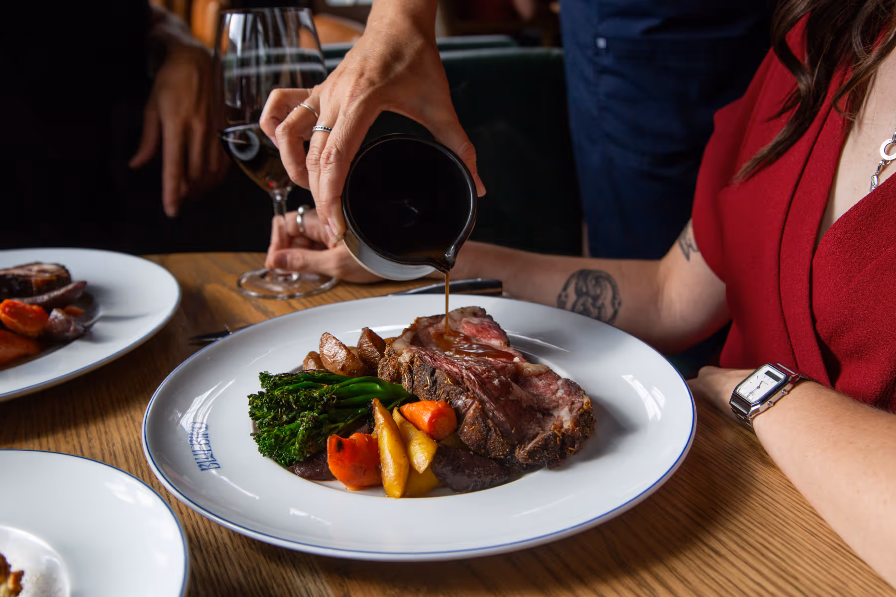 Hand pouring sauce over a steak served with roasted vegetables on a white plate at a wooden table.