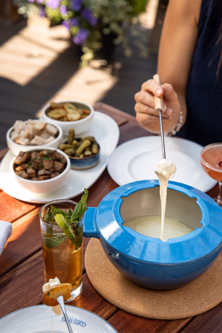 Person dipping a piece of bread into a pot of melted cheese fondue on a wooden table with drinks and side dishes.