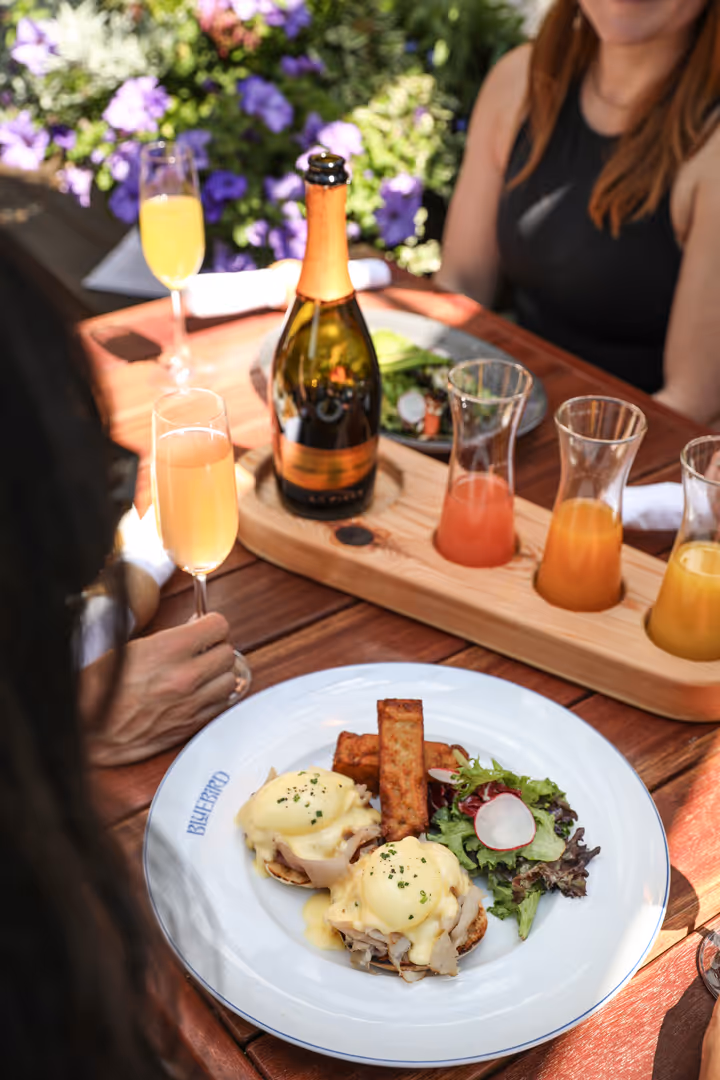 Plate with eggs Benedict, tater tots, and salad on a wooden table alongside mimosas and a bottle of sparkling wine.