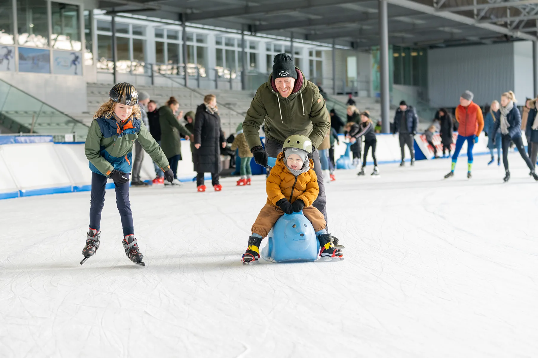 Man helpt jong kind op een blauwe zeehond-schaats op een ijsbaan met andere schaatsers op de achtergrond.