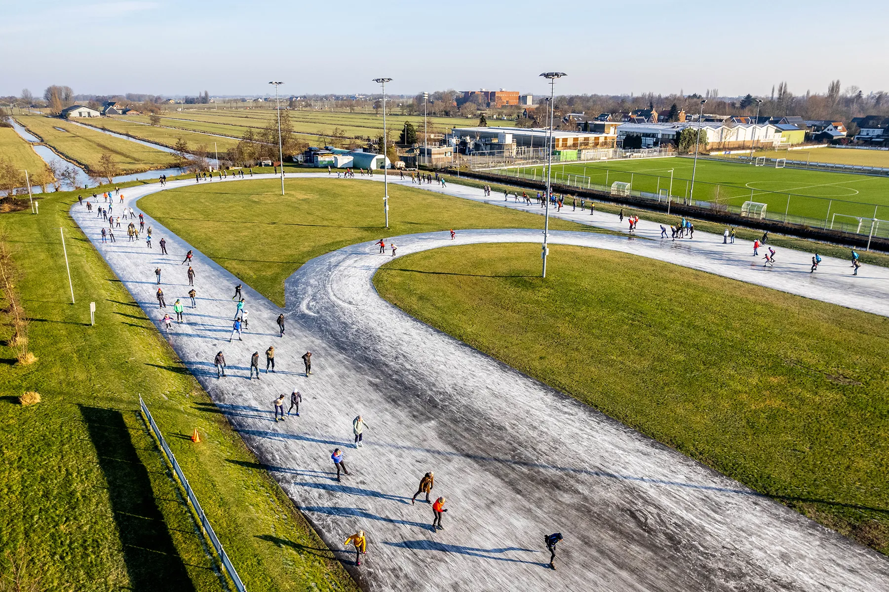 Mensen schaatsen op een gebogen natuurijsbaan omgeven door groen gras en velden onder een heldere lucht.