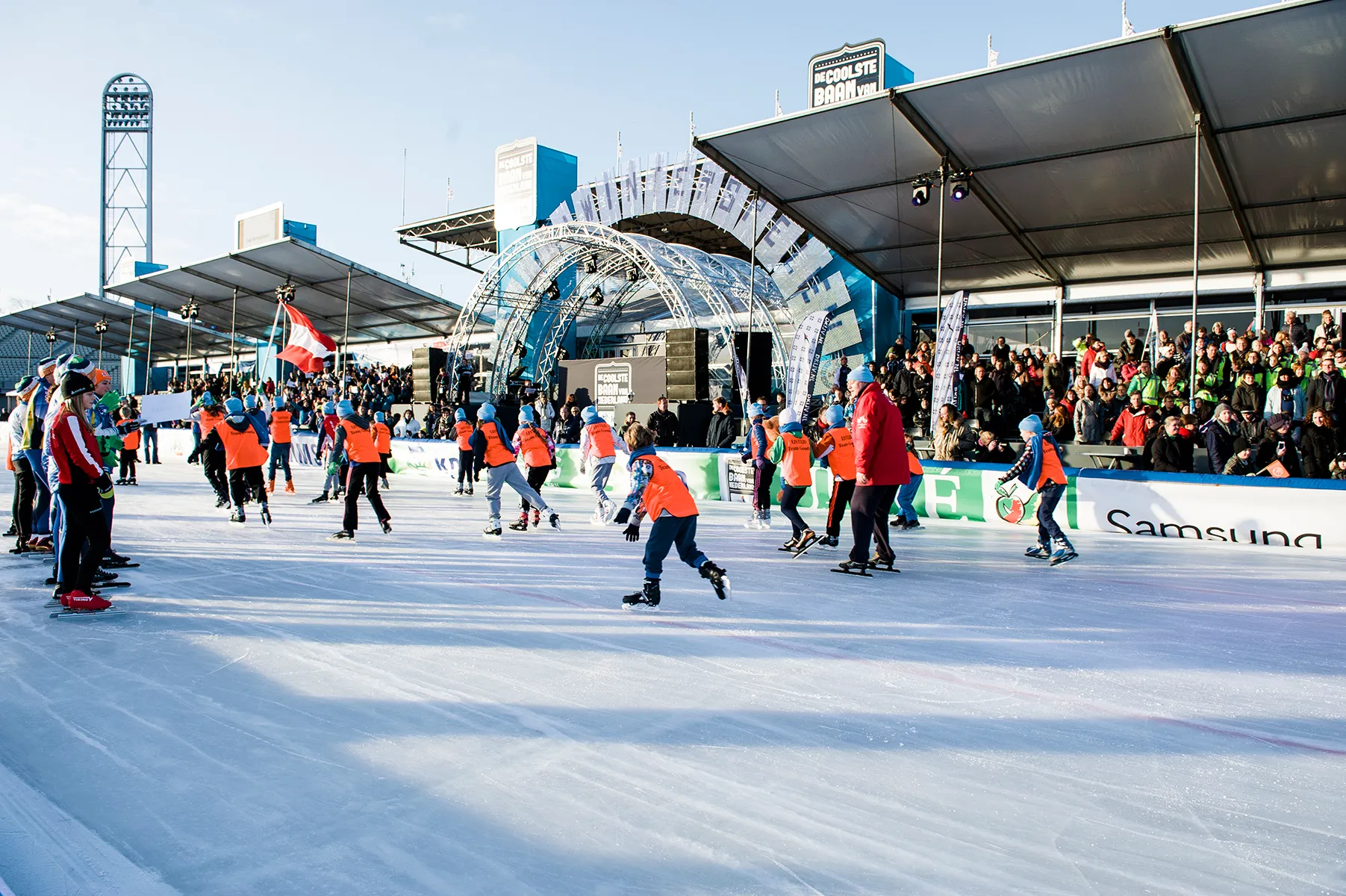 Groep kinderen met oranje hesjes schaatsen op een ijsbaan met publiek en een podium op de achtergrond.
