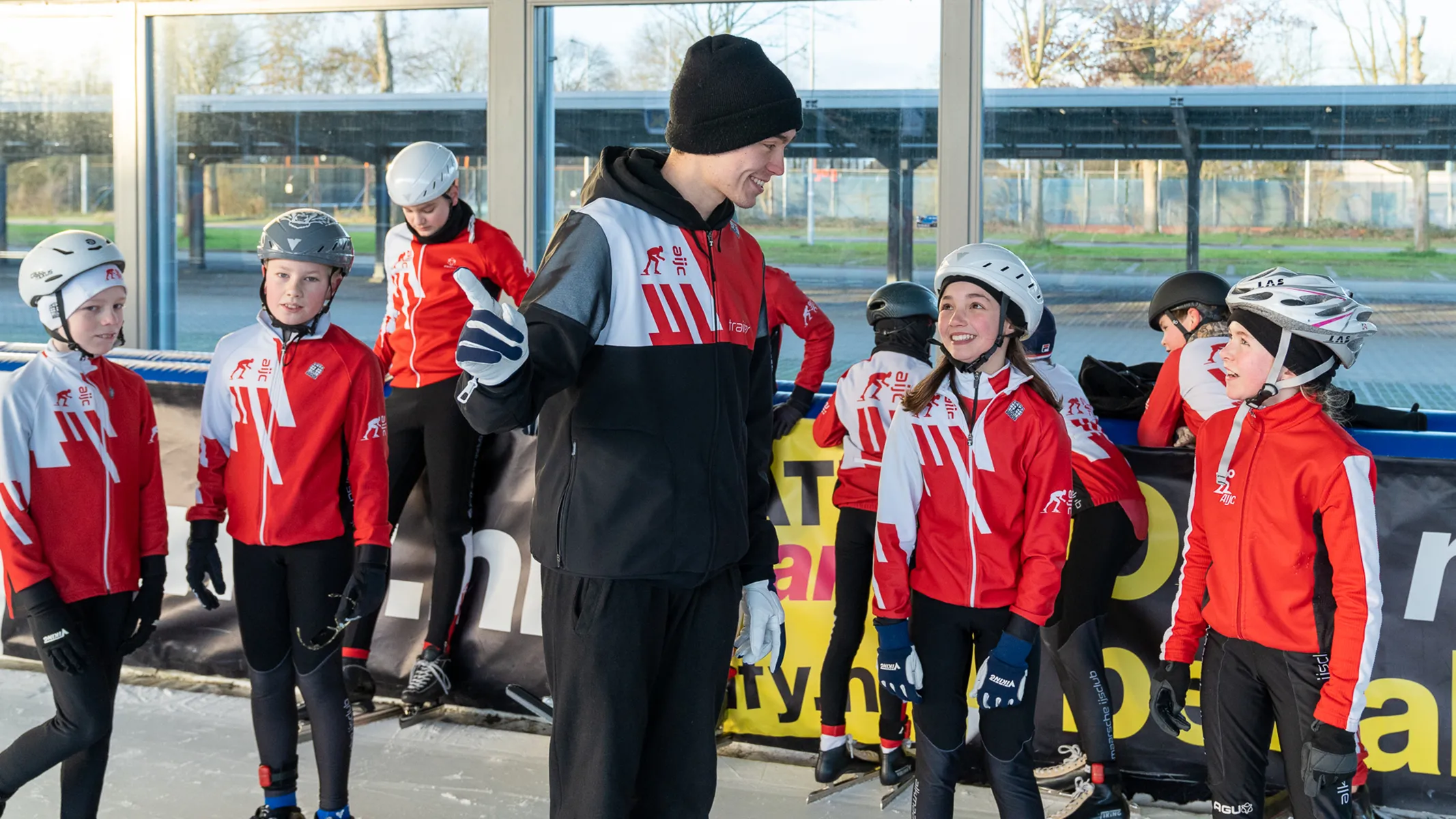 Trainer geeft instructies aan een groep jeugdige schaatsers gekleed in rood-witte trainingsjassen en helmen op een ijsbaan.