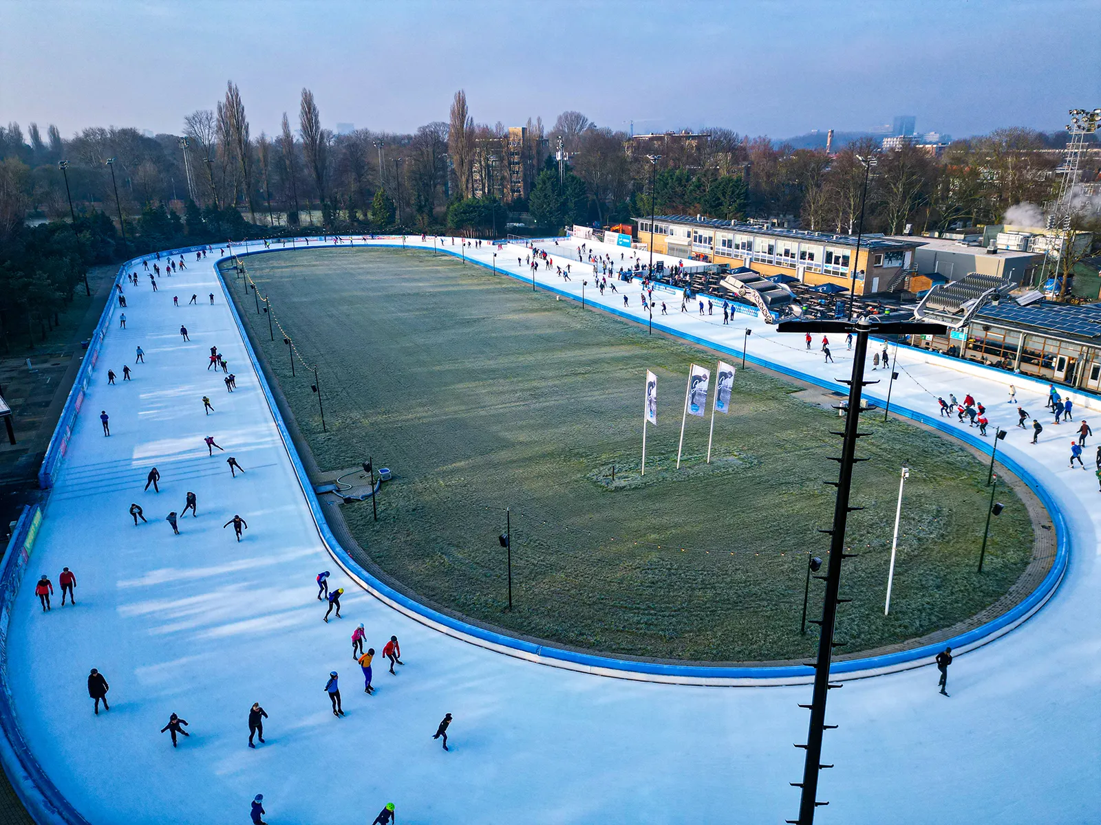 Luchtfoto van een buitenbaan voor schaatsen met meerdere mensen op het ijs en grasveld in het midden.