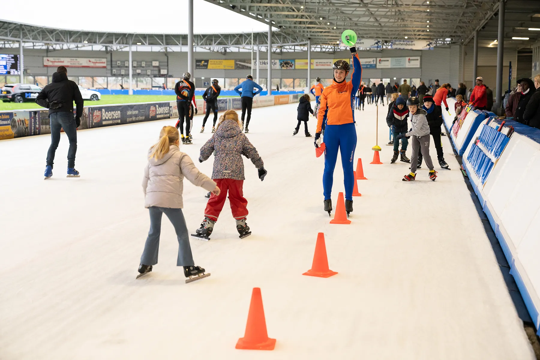 IJsbaansessie met kinderen en een instructeur in een oranje jas die een groene schijf omhoog houdt bij een rij oranje pionnen.