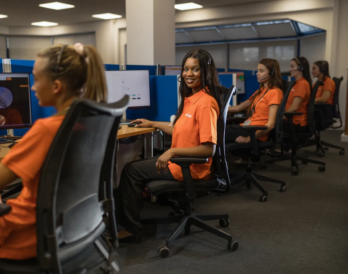 Team members in call center wearing orange uniforms seated at workstations with computer monitors, headsets, and office chairs.