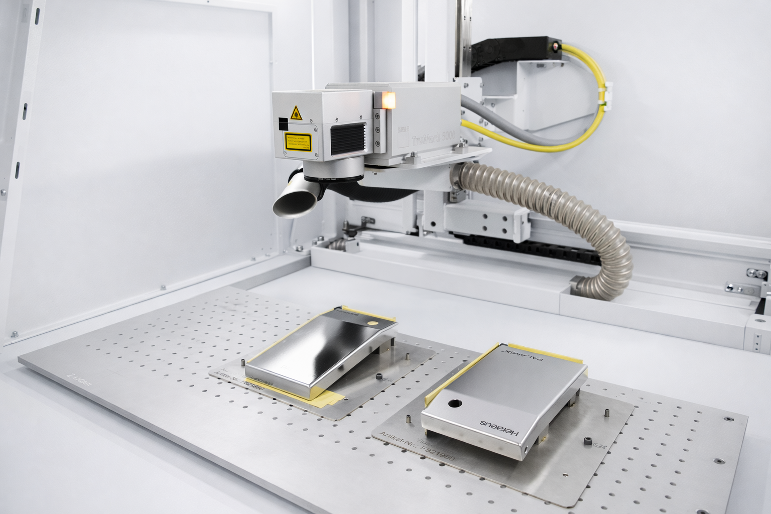 Industrial laser machine analyzing two metallic smartphone casings on a perforated metal table.