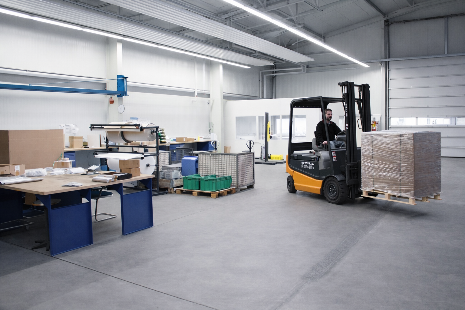 Worker operating forklift carrying pallet wrapped boxes inside a spacious warehouse with packaging materials and work tables.