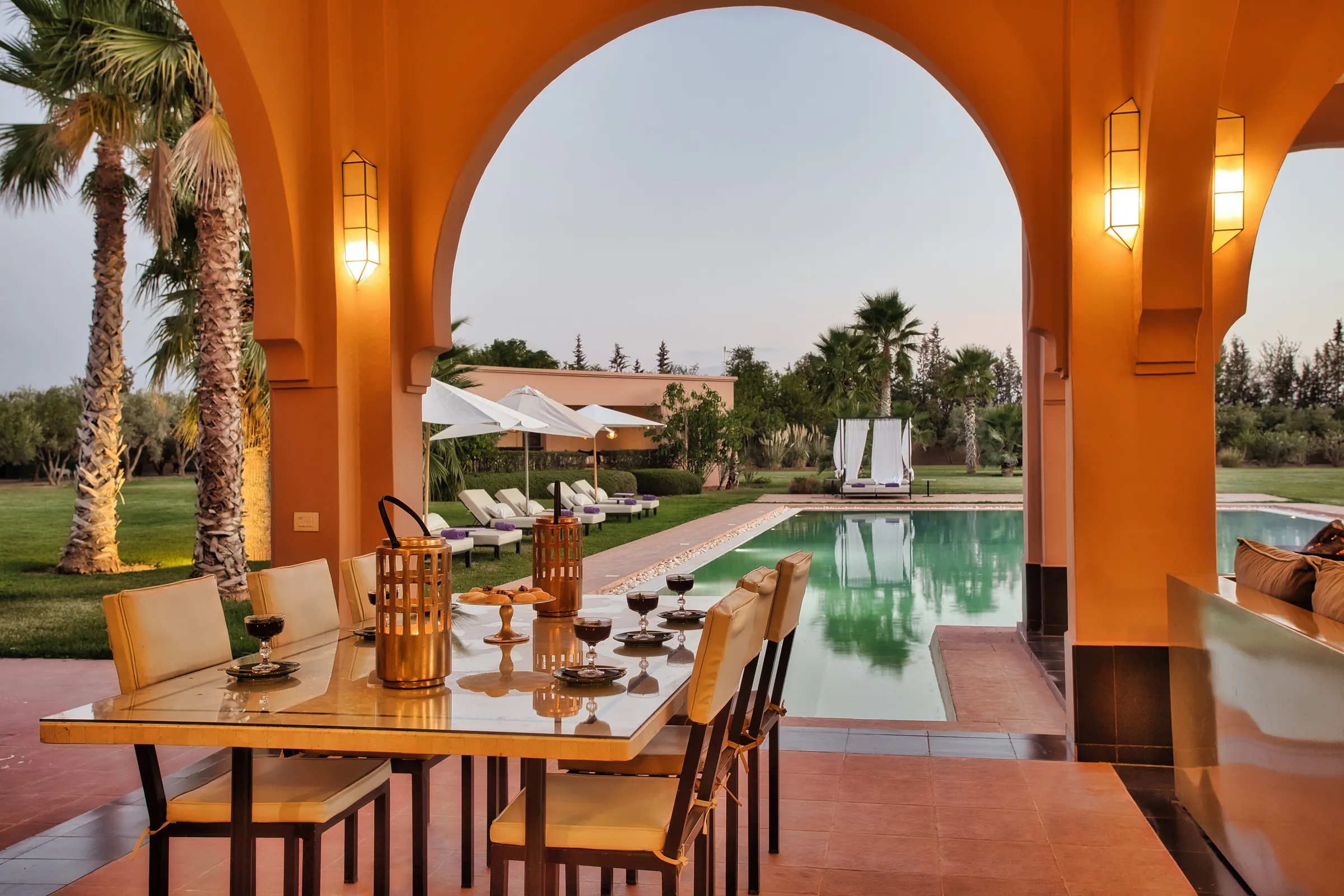 Villa Theia, private villa rental on the Route de l'Ourika, Marrakech: outdoor dining table set under the arches overlooking the pool at dusk
