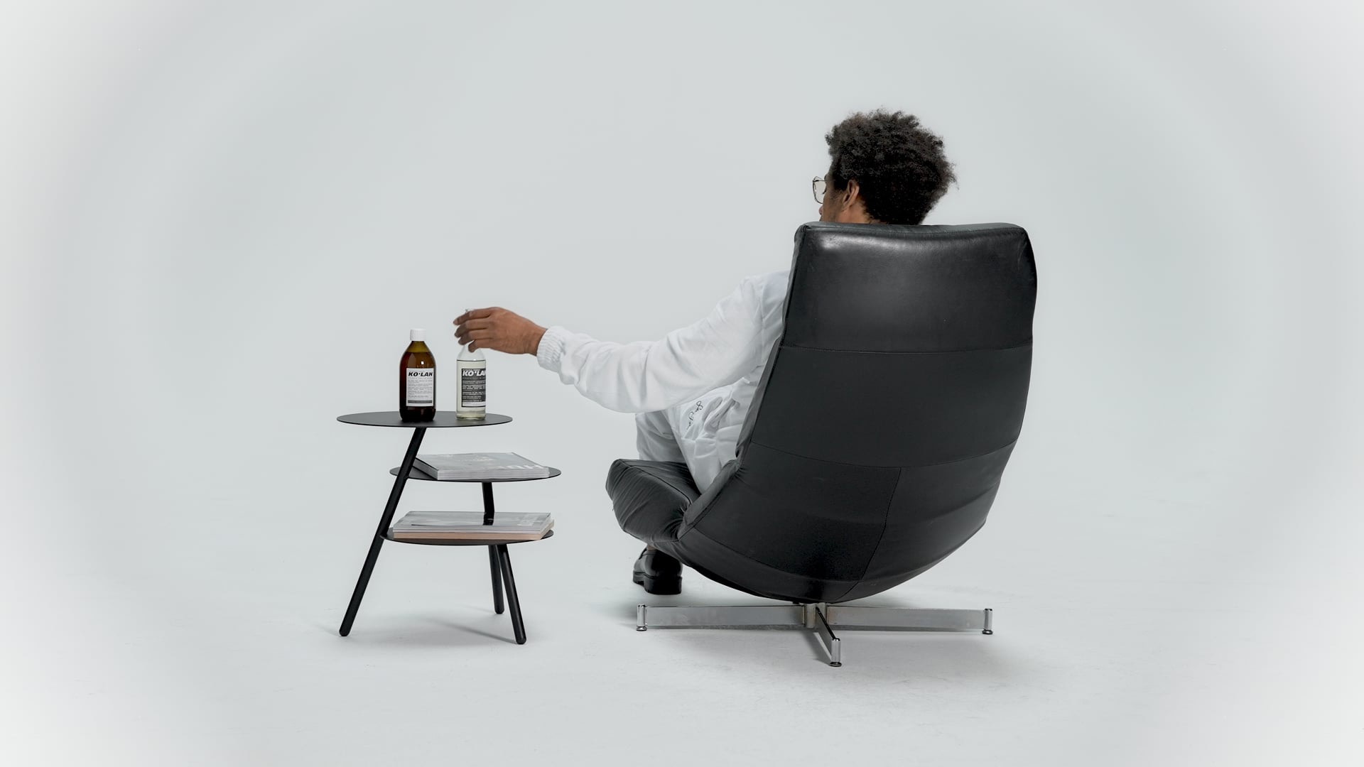 Person sitzt entspannt in einem schwarzen Loungesessel vor weißem Hintergrund. Daneben stehen auf einem Beistelltisch zwei Flaschen und Magazine in minimalistischem Studio-Setting.