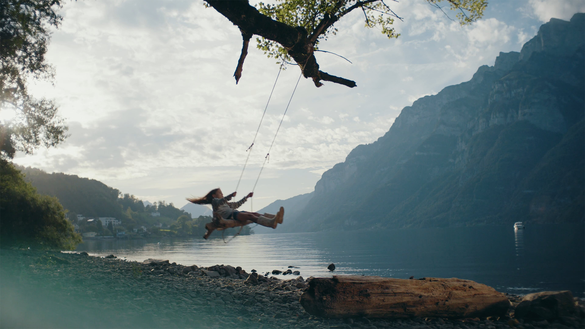 Eine Person schwingt sich an einem Seil unter einem Baum über einen ruhigen See, im Hintergrund sind Berge zu sehen