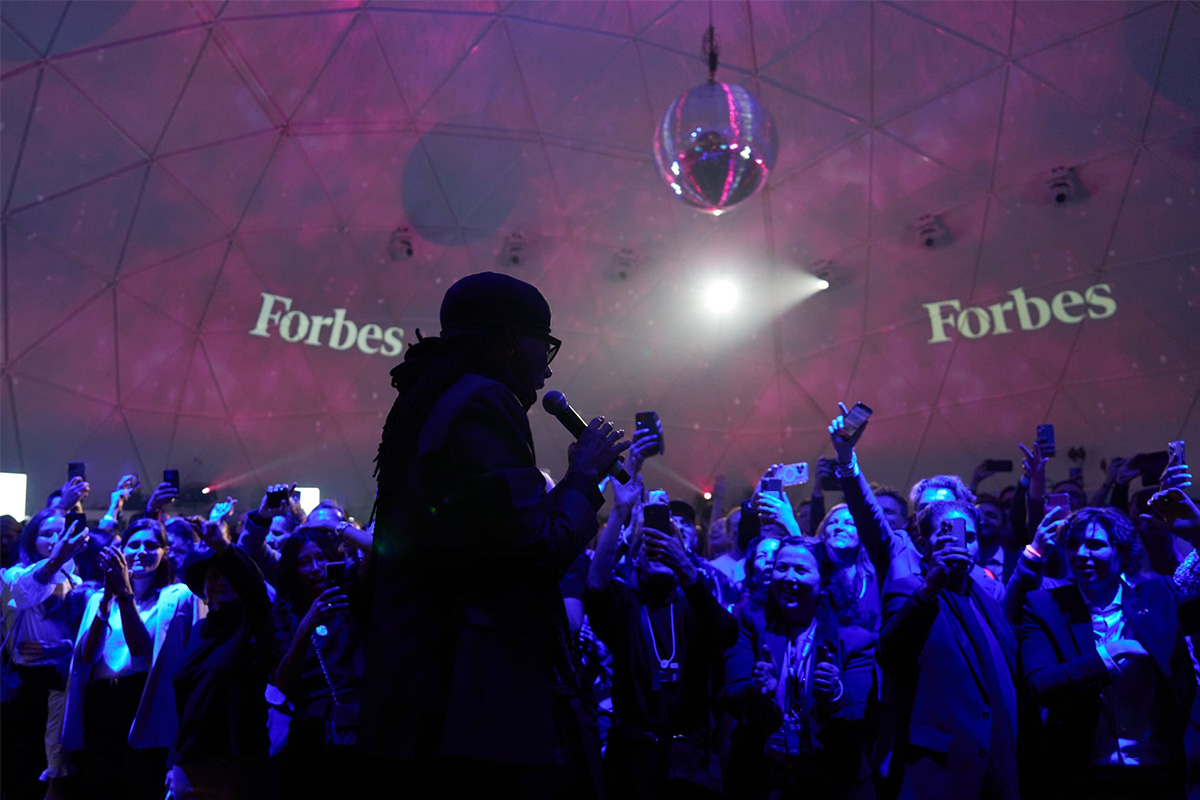 Sänger mit Mikrofon performt vor Publikum unter Disco-Kugel, „Forbes“-Logo im Hintergrund