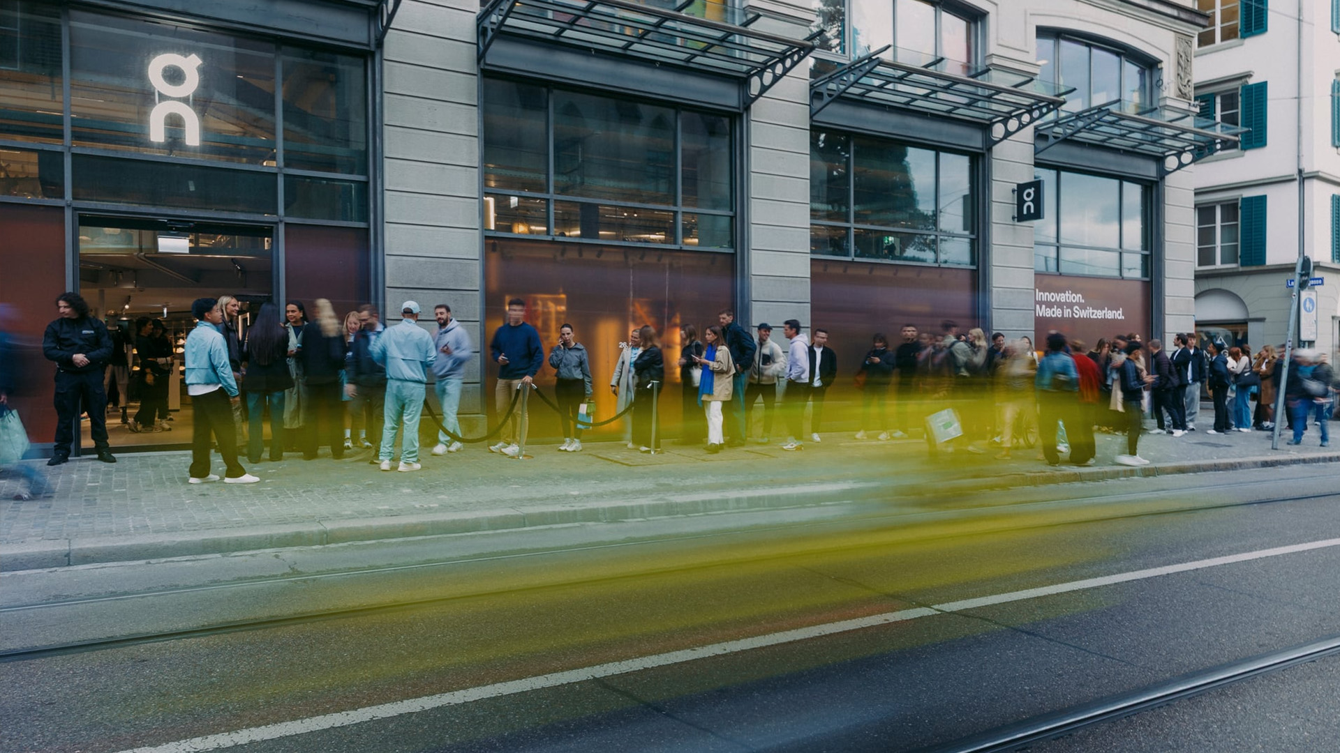 Warteschlange vor einem modernen Store in urbaner Umgebung, während vorbeifahrende Fahrzeuge als Licht- und Bewegungsstreifen im Vordergrund sichtbar sind.