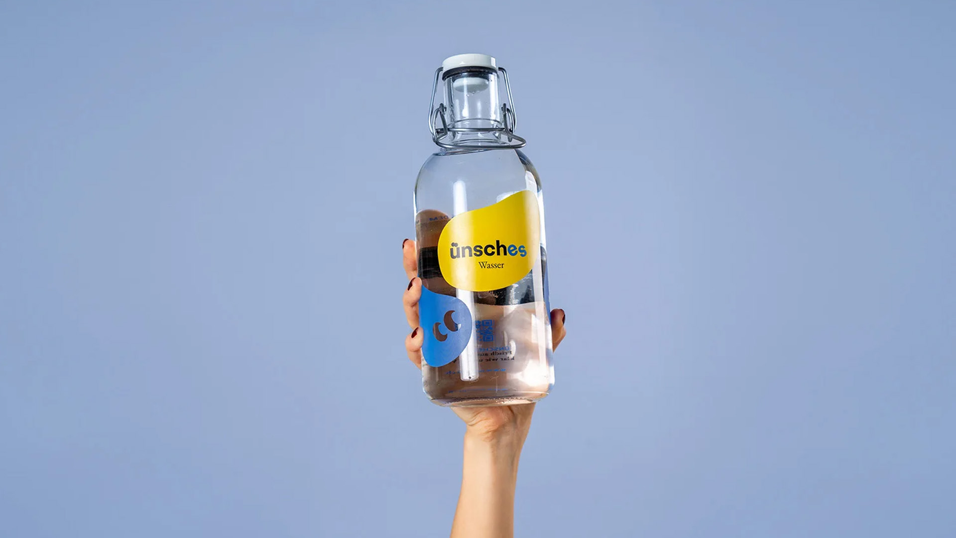 Hand hält eine wiederverwendbare Glasflasche mit „Ünsches Wasser“-Branding vor klarem, blauem Himmel.