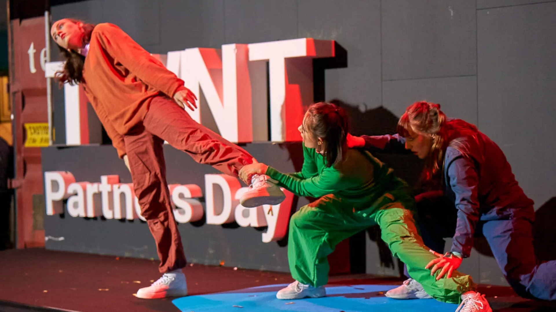 Drei Tänzerinnen in farbigen Overalls performen eine dynamische Choreografie auf einer Bühne vor beleuchteten Event-Schriftzügen.