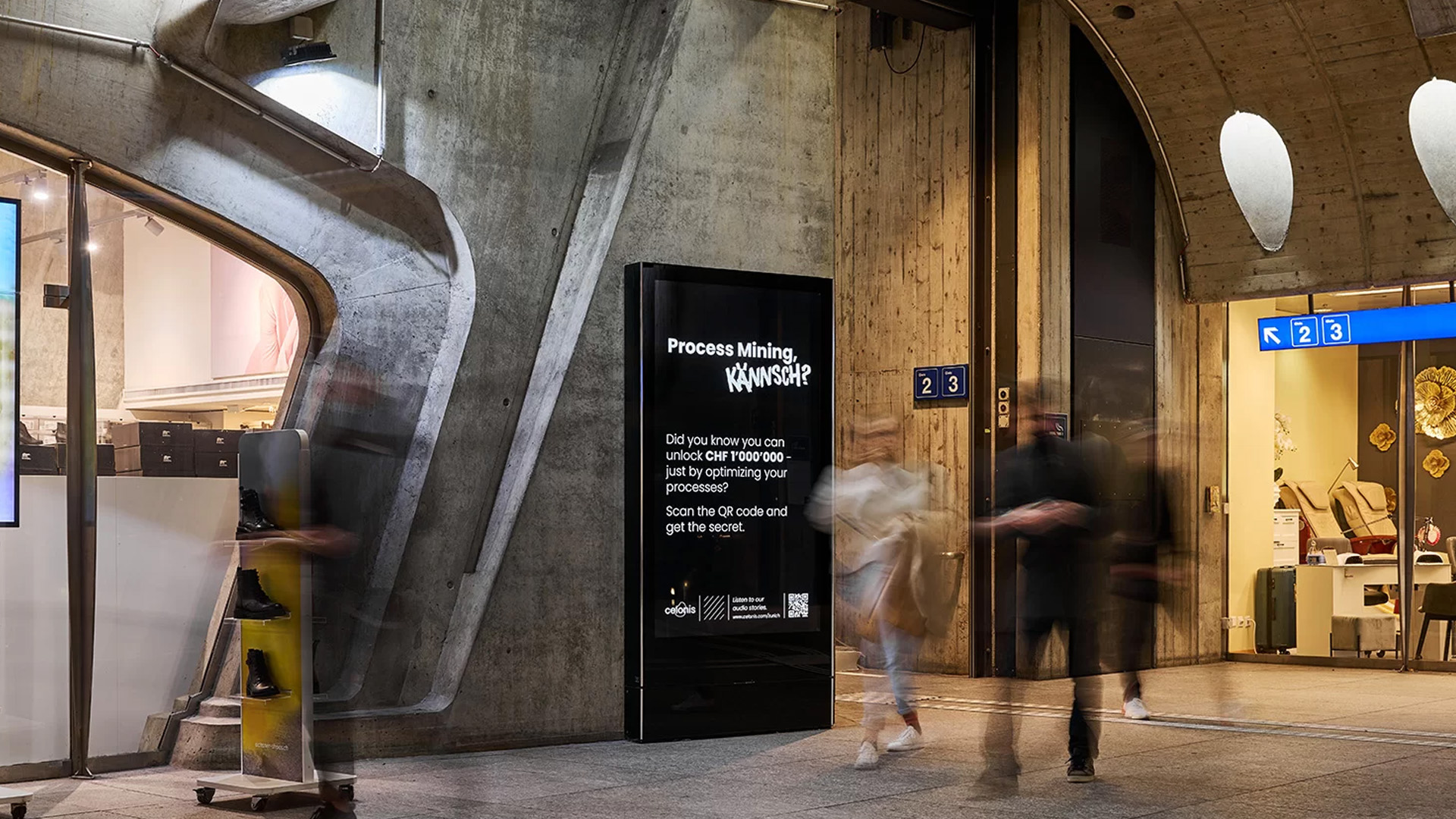 Digitale Werbefläche in einer modernen Bahnhofspassage mit vorbeieilenden Passantinnen und Passanten vor markanter Betonarchitektur.