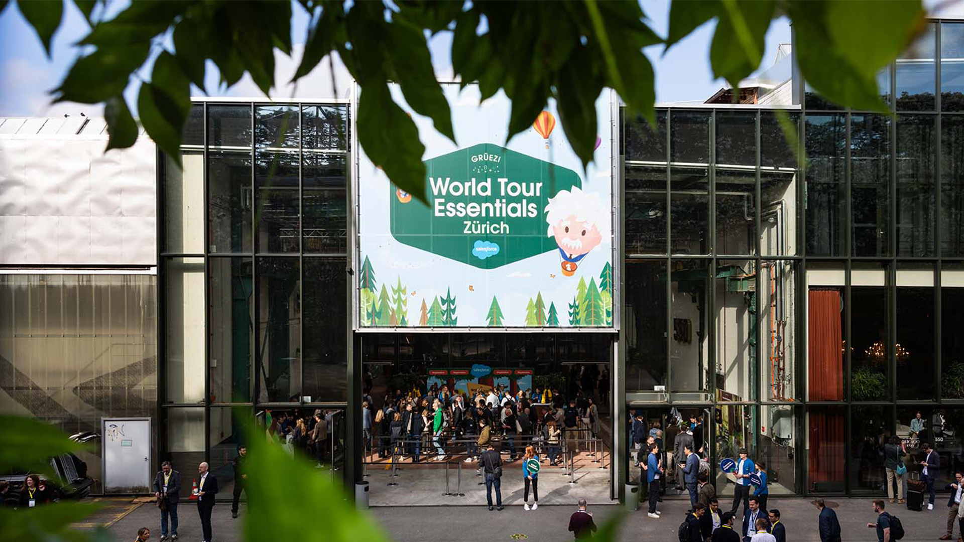 Aussenansicht der Salesforce World Tour Essentials Zürich mit grossflächigem Branding am Eingang, Besuchenden vor der Location und grünen Blättern im Vordergrund.