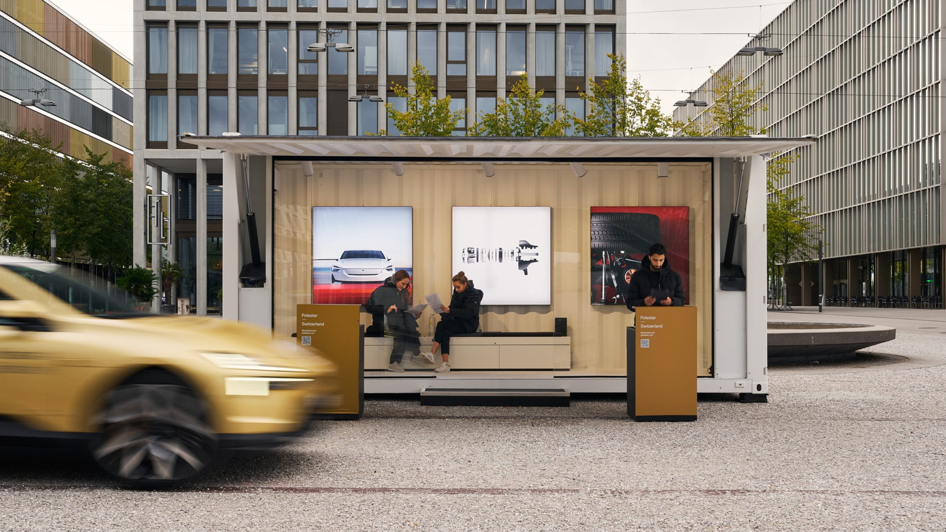 Offener Ausstellungscontainer von Polestar auf einem urbanen Platz, mit grossformatigen Fahrzeugmotiven im Innenraum, Besuchenden im Container und einem vorbeifahrenden Auto im Vordergrund.