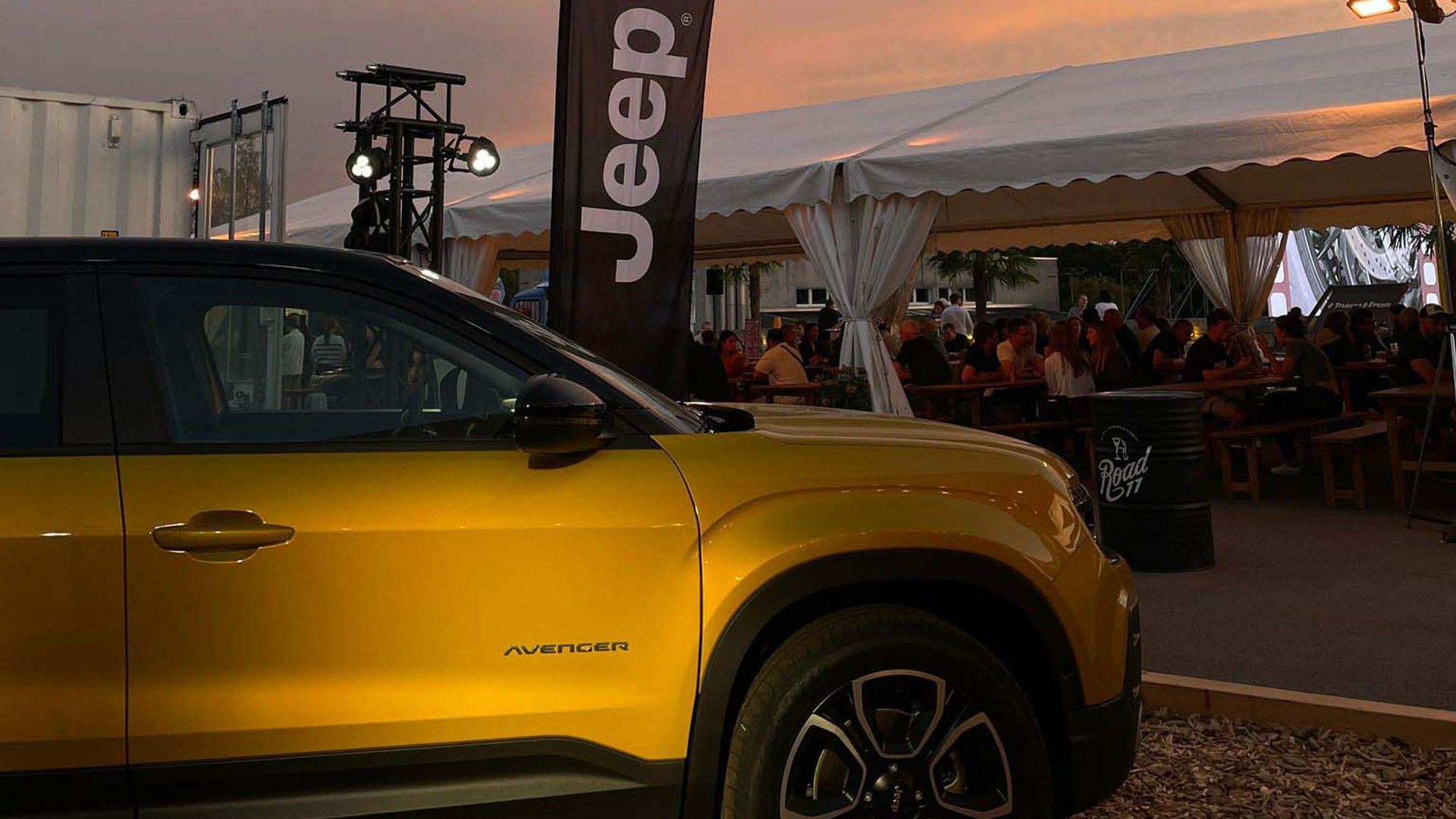 Gelber Jeep Avenger vor einem beleuchteten Eventzelt bei Abendstimmung, während Gäste im Hintergrund an Tischen zusammensitzen und die Veranstaltung geniessen.