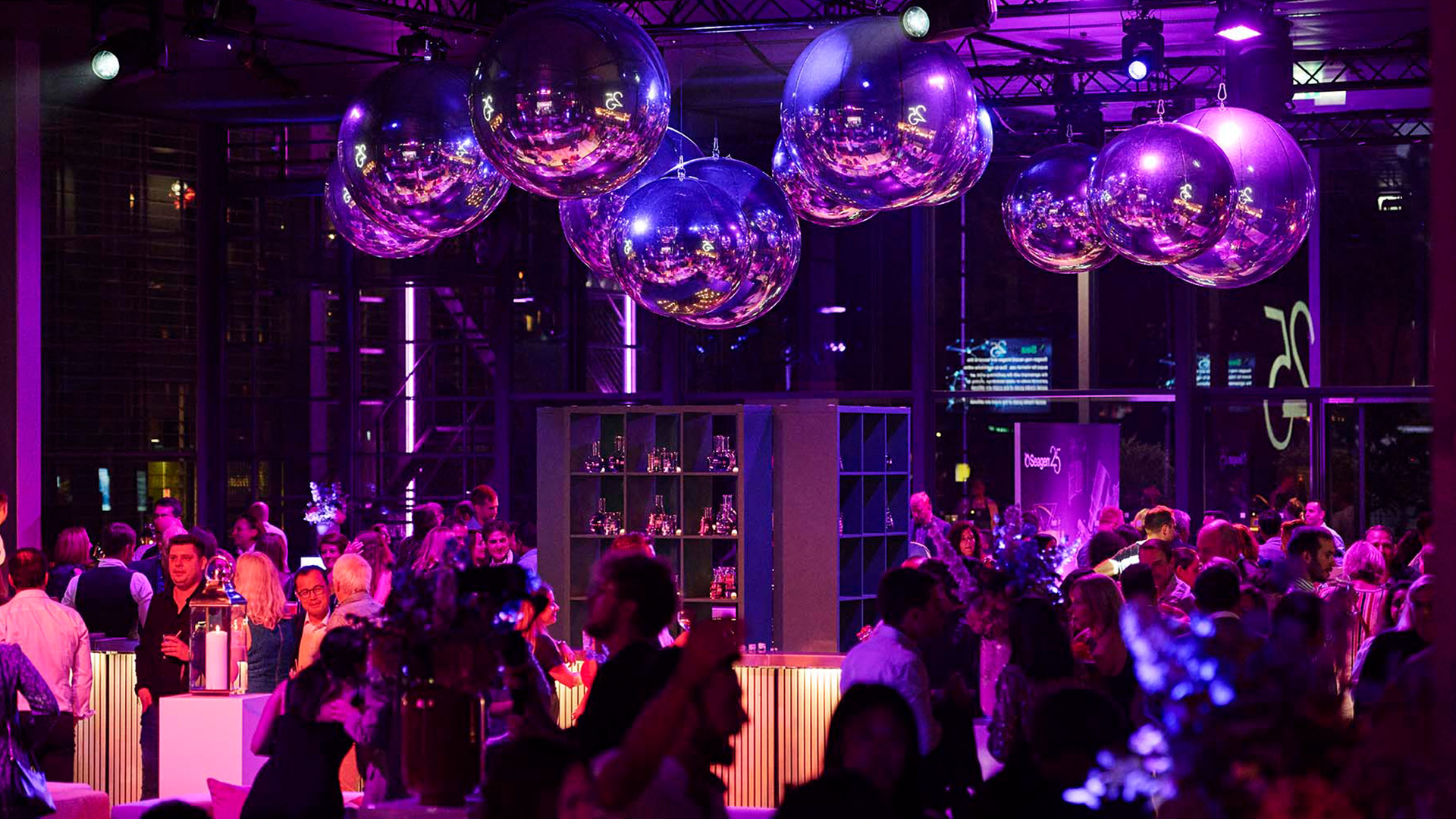 Festliche Eventlocation mit Gästen, stimmungsvoller violetter Beleuchtung und grossen verspiegelten Kugeln als Deckendekoration.