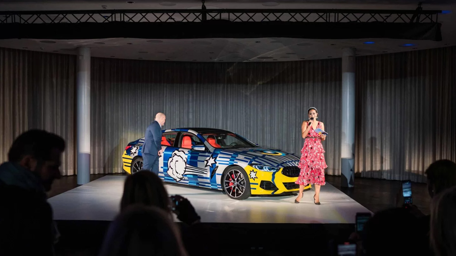 A strikingly designed BMW is being presented on stage at an event. A male and female presenter, each holding a microphone, stand next to the vehicle while the audience watches the presentation.