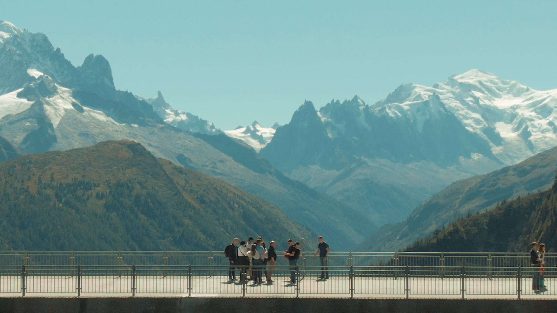 Menschen auf einer Aussichtsplattform vor schneebedeckten Alpenbergen und einem weiten Bergtal bei klarem Himmel.