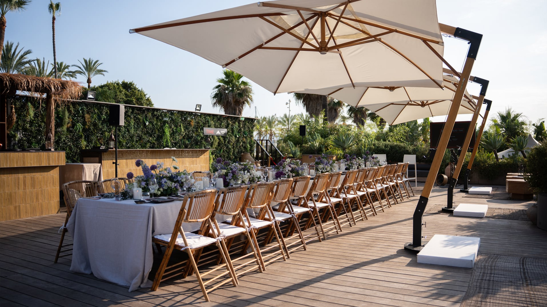 Festlich gedeckte Tafel auf einer Terrasse mit Sonnenschirmen, Blumenarrangements und mediterraner Gartenkulisse.