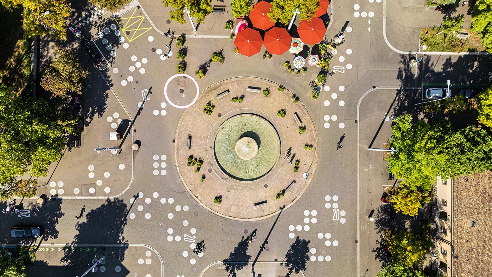 Luftaufnahme eines begrünten Stadtplatzes mit rundem Brunnen, roten Sonnenschirmen und grafischen Markierungen auf den Wegen.