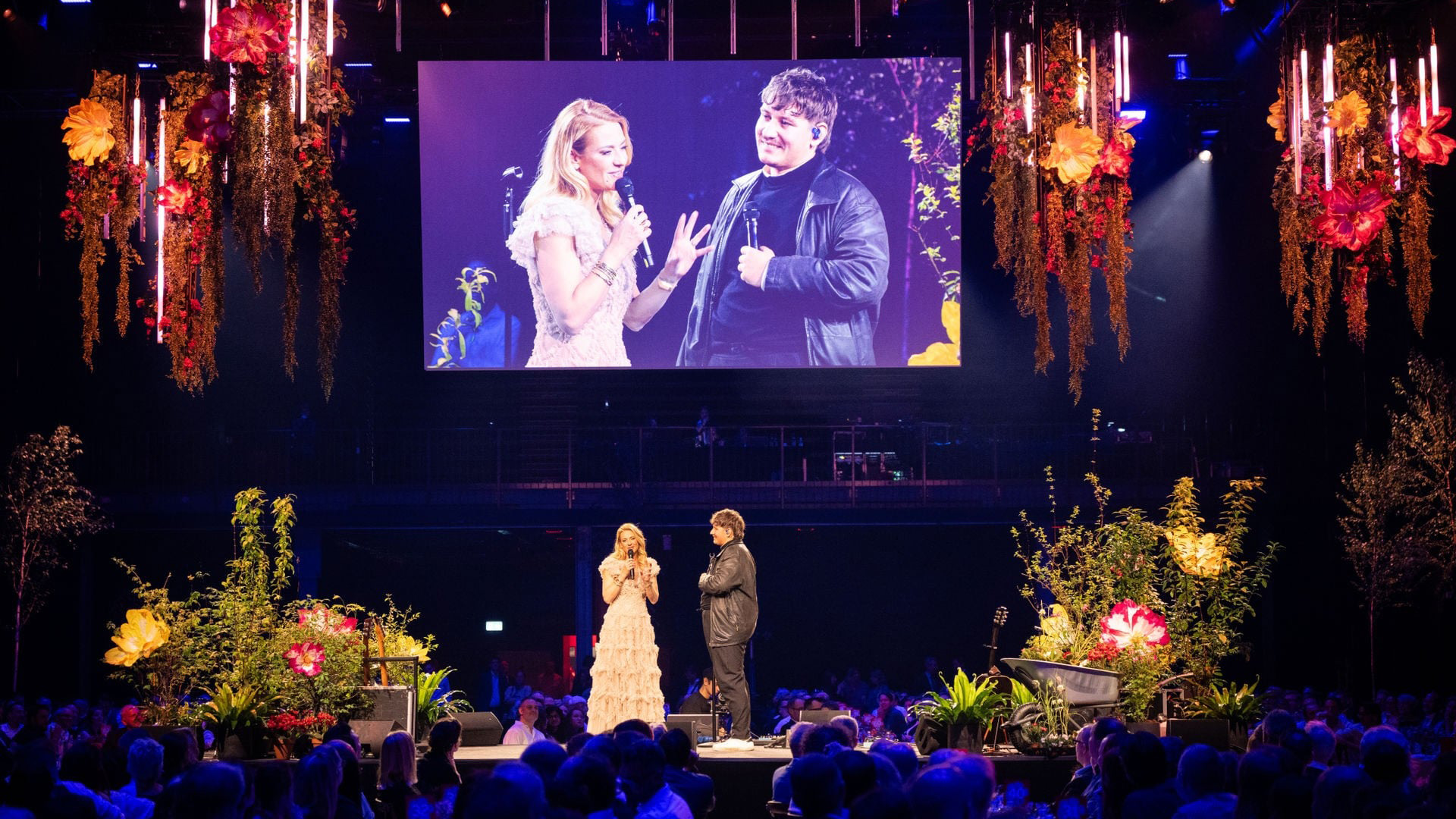 Bühnengespräch auf floraler Eventbühne vor Publikum, mit grosser LED-Leinwand und dekorativen Blumeninstallationen.