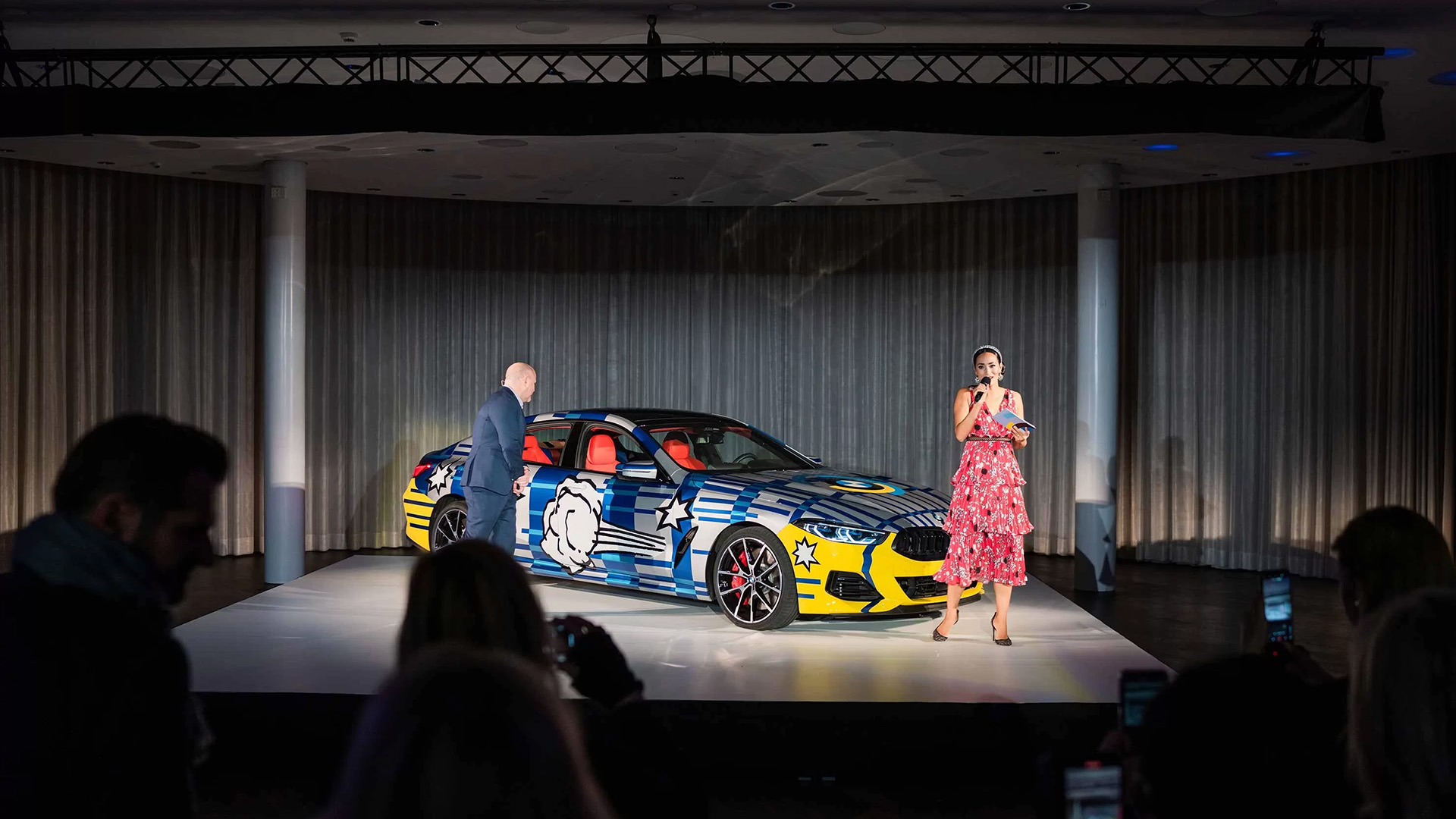 Präsentation eines auffällig gestalteten BMW auf einer Bühne bei einem Event. Neben dem Fahrzeug stehen ein Moderator und eine Moderatorin mit Mikrofon, während das Publikum die Vorstellung verfolgt.
