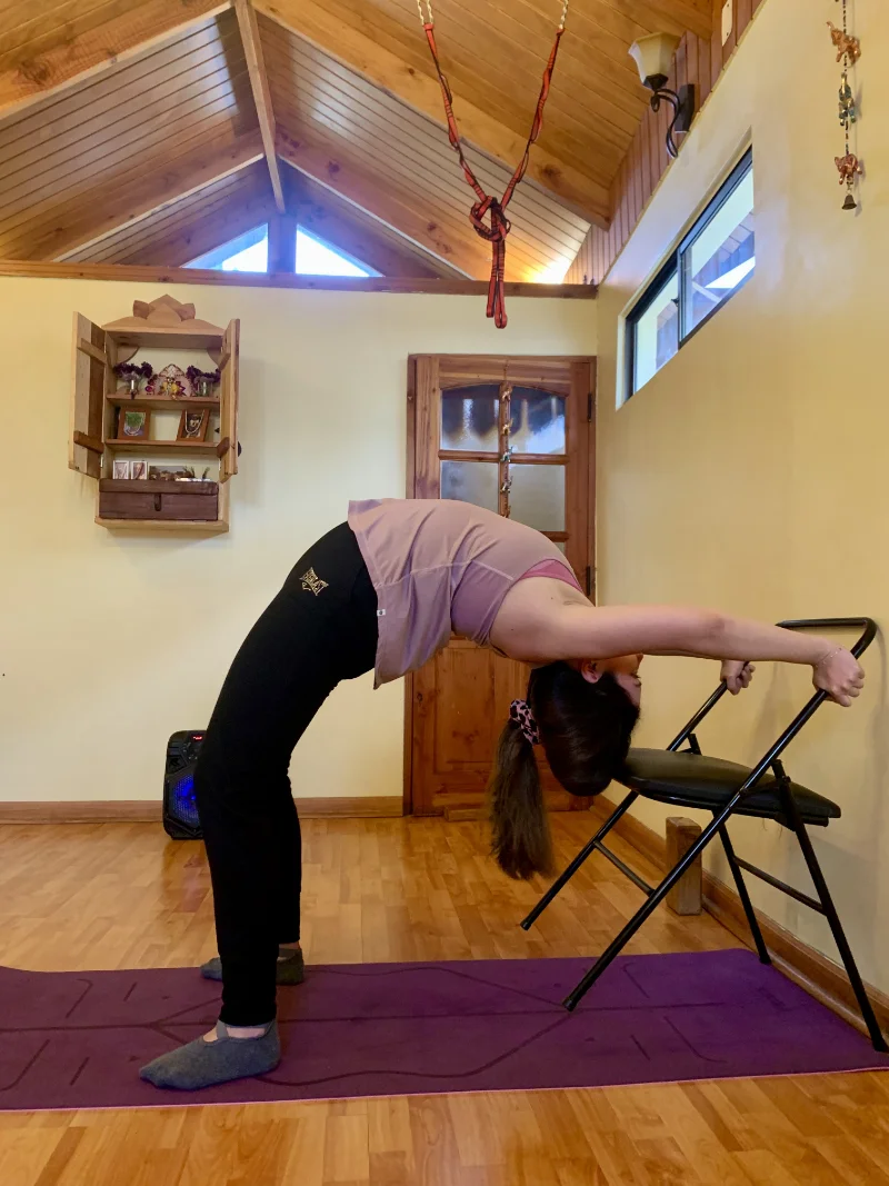 Woman performing a yoga backbend using a chair in a room with wooden floor and ceiling.
