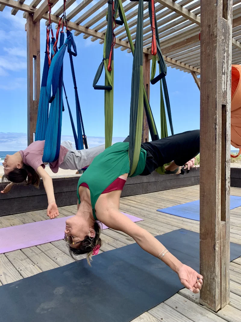 Two women practicing aerial yoga suspended upside down in fabric hammocks under a wooden pergola near a beach.