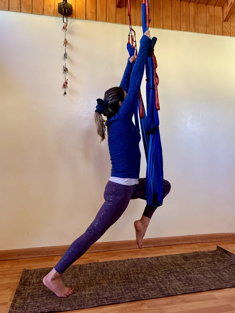 Woman practicing aerial yoga indoors using blue aerial hammock while in a lunge pose with one foot on the floor and the other bent forward.