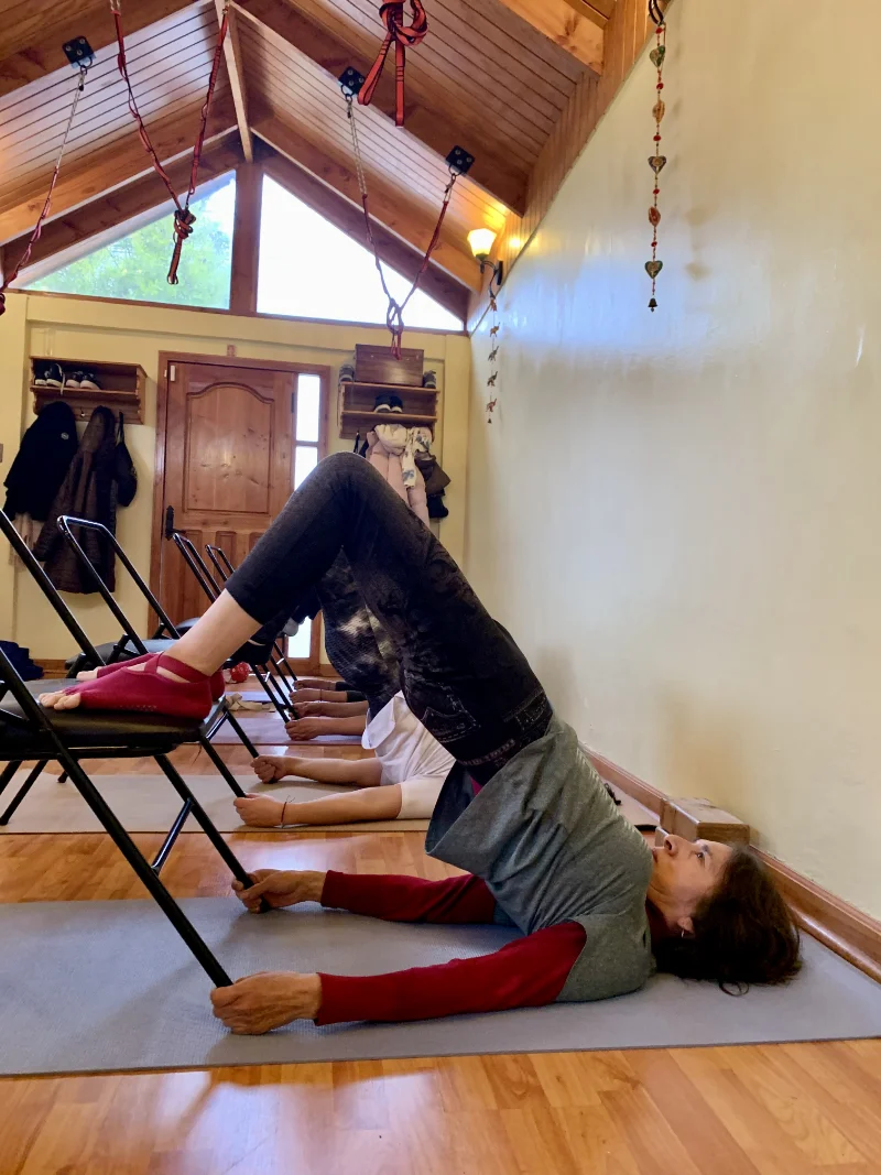 Two women in a yoga class doing bridge pose with feet on chairs inside a room with wooden ceiling and coats hanging by the door.