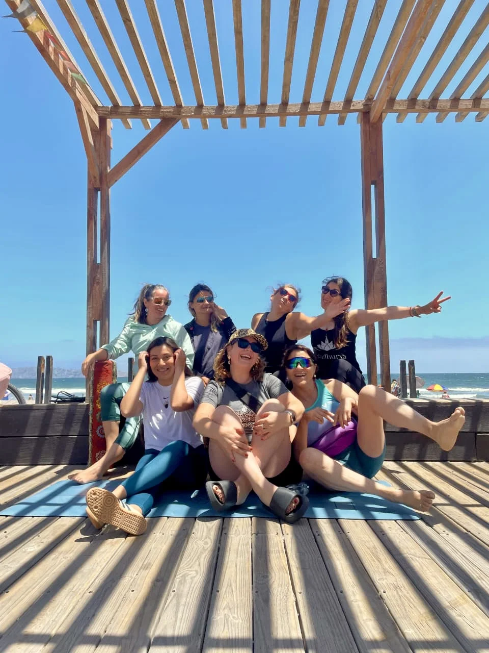 Group of six women sitting and posing playfully under a wooden pergola on a sunny beach boardwalk.