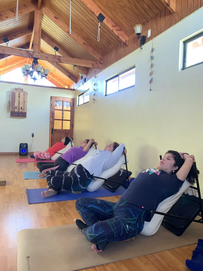 Four women practicing seated yoga stretches with back support in a wooden-floored room with high wooden ceiling and windows.